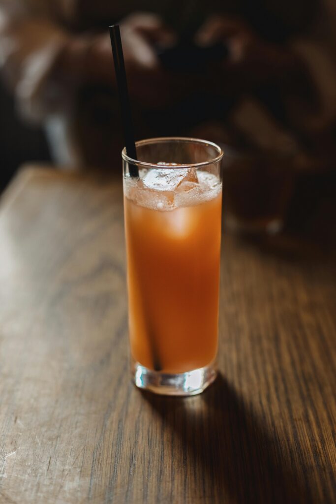 A glass of orange iced tea with ice cubes and a straw on a dimly lit wooden table.
